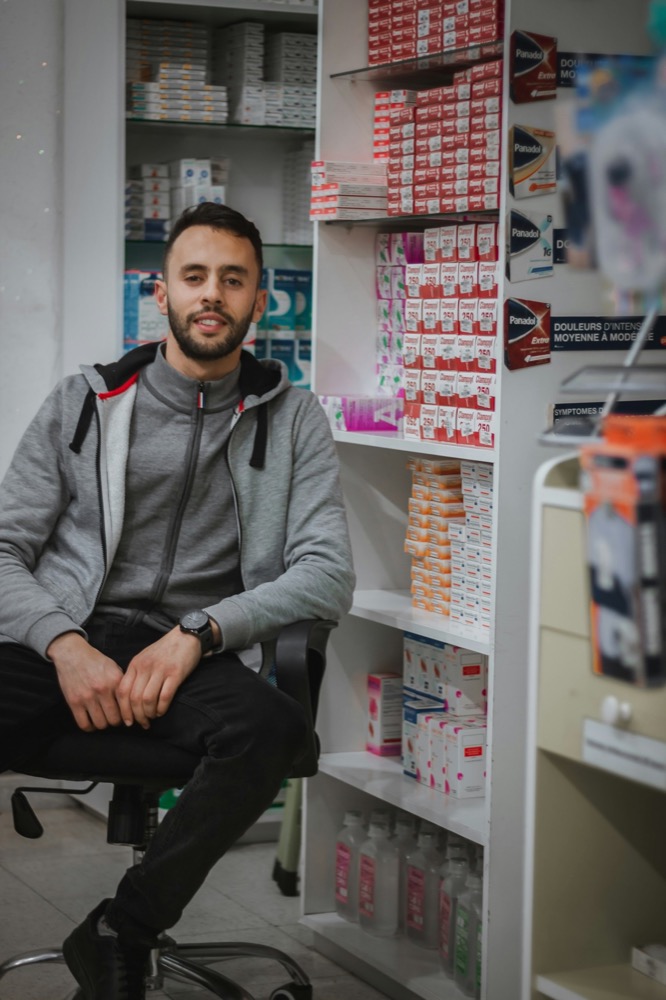 A young man with a beard sits in a chair in front of shelves filled with medication boxes in a pharmacy.
