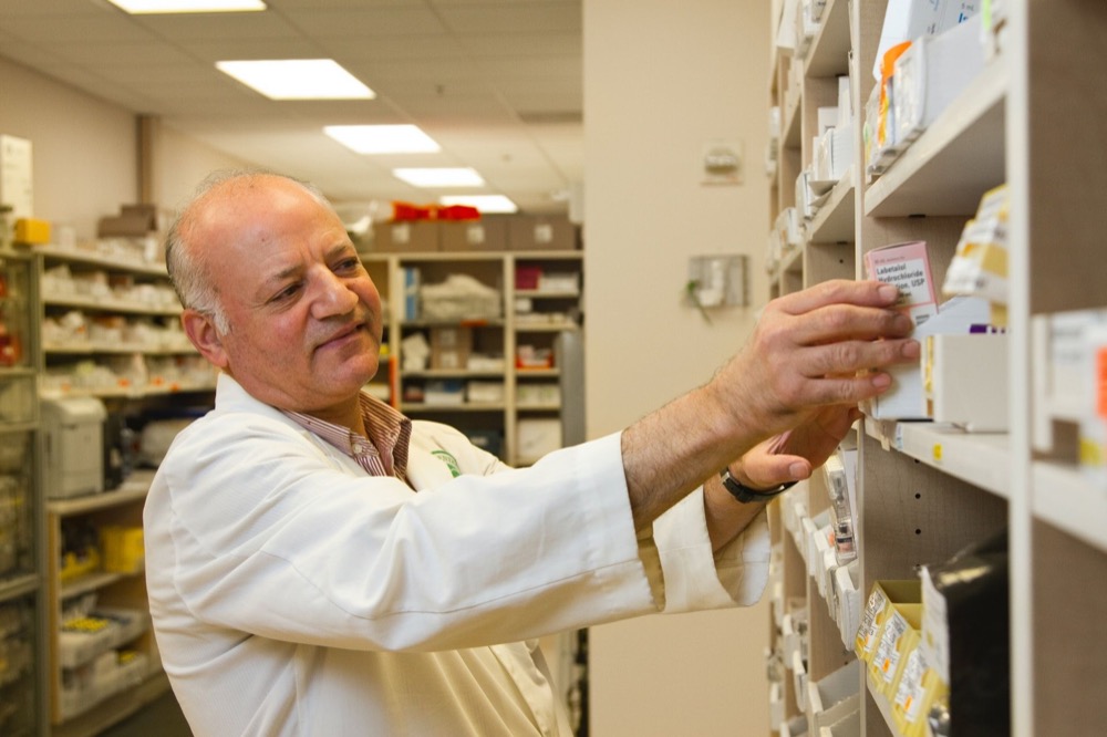 A male pharmacist in a white lab coat reaches for medication on a shelf in a pharmacy.